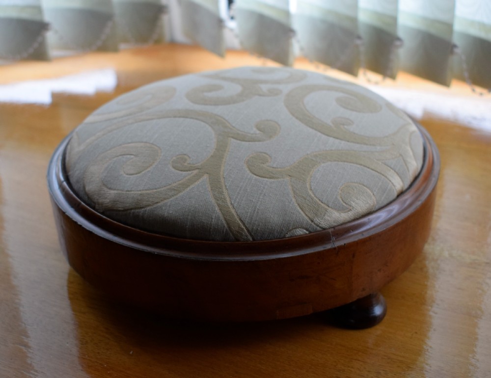 A circular Victorian footstool with a polished brown walnut frame, small turned feet, and a padded cream textile top featuring a scroll motif.