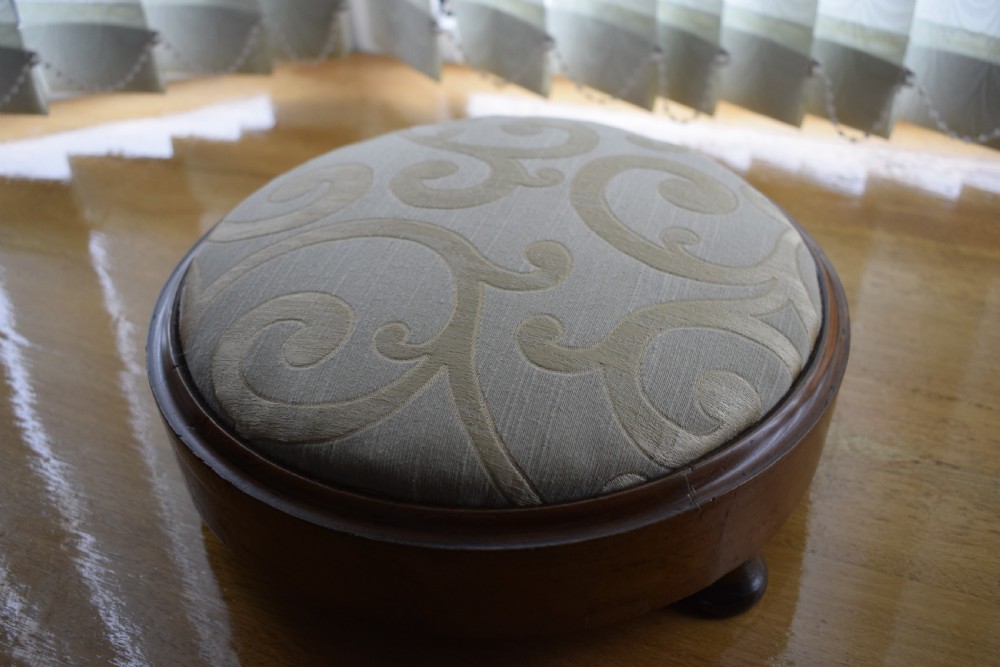 A circular Victorian footstool with a polished brown walnut frame, small turned feet, and a padded cream textile top featuring a scroll motif. View III