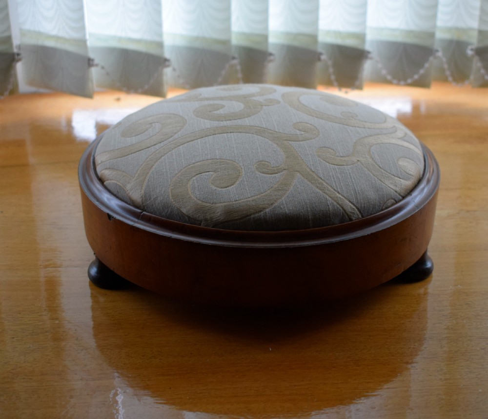 A circular Victorian footstool with a polished brown walnut frame, small turned feet, and a padded cream textile top featuring a scroll motif. View IV