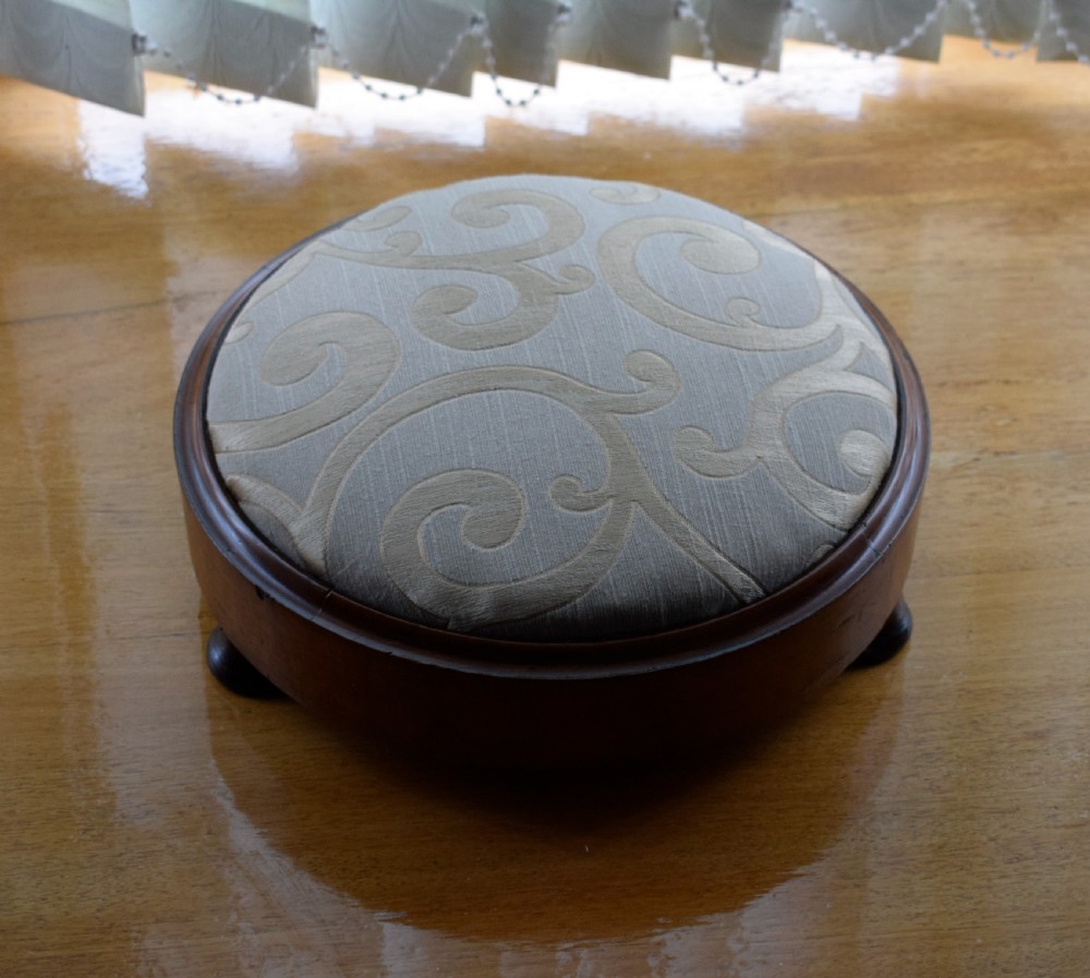 A circular Victorian footstool with a polished brown walnut frame, small turned feet, and a padded cream textile top featuring a scroll motif. View V