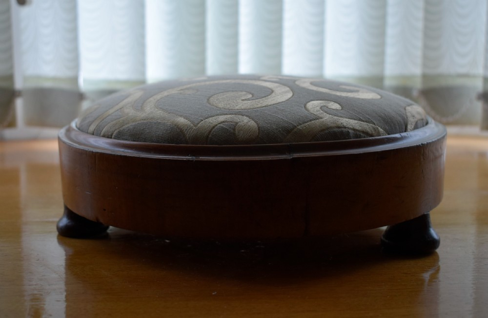 A circular Victorian footstool with a polished brown walnut frame, small turned feet, and a padded cream textile top featuring a scroll motif. View VI