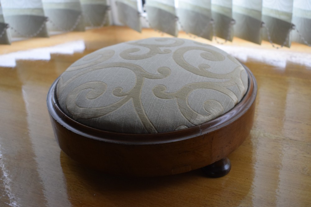 A circular Victorian footstool with a polished brown walnut frame, small turned feet, and a padded cream textile top featuring a scroll motif. View VII