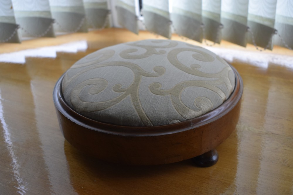 A circular Victorian footstool with a polished brown walnut frame, small turned feet, and a padded cream textile top featuring a scroll motif. View VIII