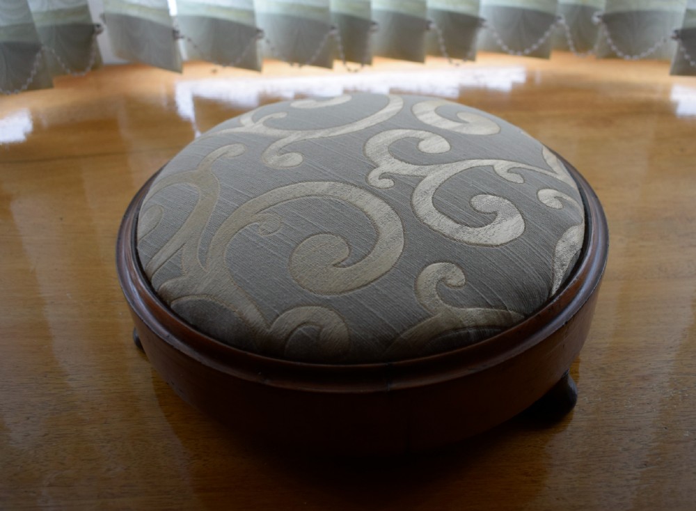 A circular Victorian footstool with a polished brown walnut frame, small turned feet, and a padded cream textile top featuring a scroll motif. View IX