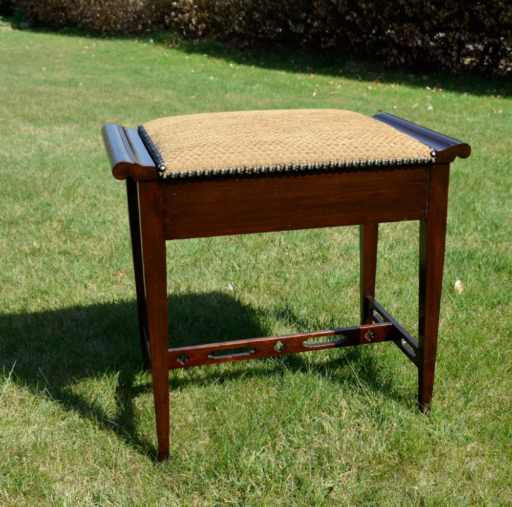 An Arts and Crafts dark wood piano stool featuring a textured tan upholstered seat with brass studding, raised side handles, tapered legs, and a pierced decorative lower stretcher.