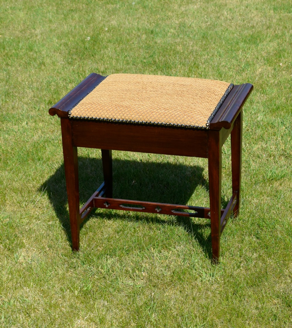 An Arts and Crafts dark wood piano stool featuring a textured tan upholstered seat with brass studding, raised side handles, tapered legs, and a pierced decorative lower stretcher. View II