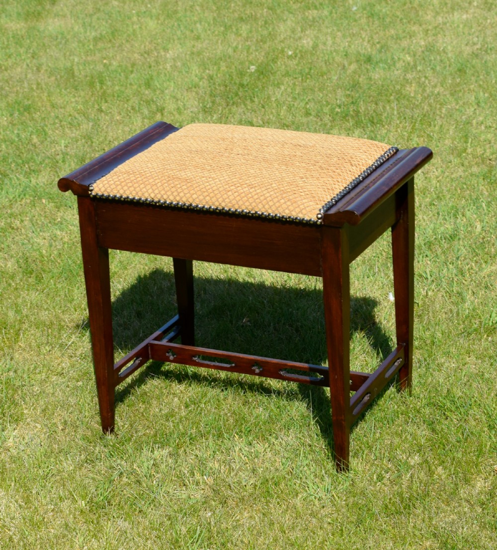 An Arts and Crafts dark wood piano stool featuring a textured tan upholstered seat with brass studding, raised side handles, tapered legs, and a pierced decorative lower stretcher. View III