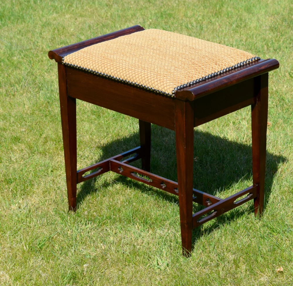An Arts and Crafts dark wood piano stool featuring a textured tan upholstered seat with brass studding, raised side handles, tapered legs, and a pierced decorative lower stretcher. View IV