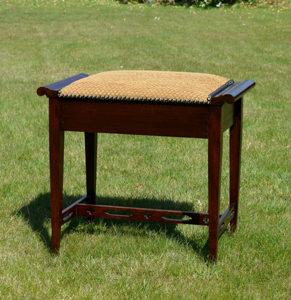 An Arts and Crafts dark wood piano stool featuring a textured tan upholstered seat with brass studding, raised side handles, tapered legs, and a pierced decorative lower stretcher. View V