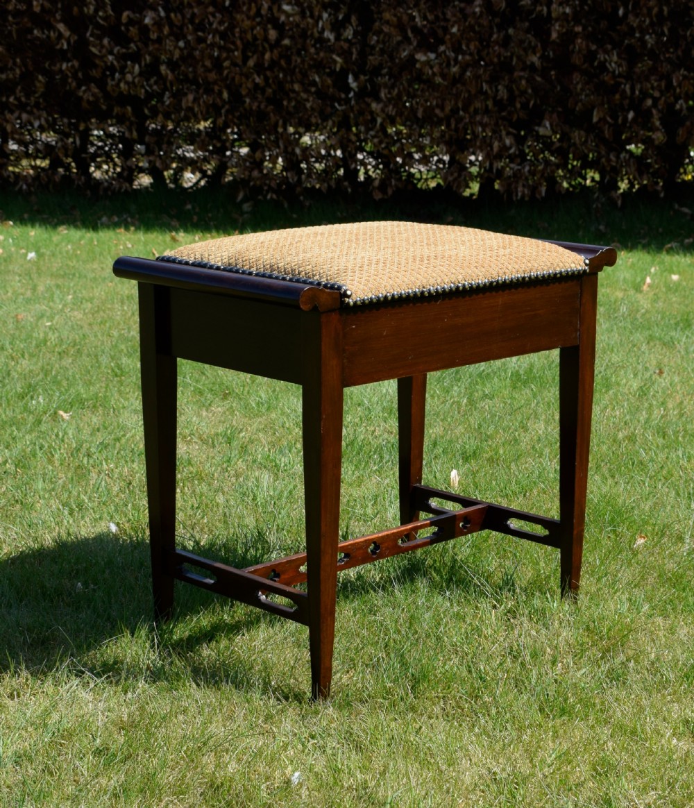 An Arts and Crafts dark wood piano stool featuring a textured tan upholstered seat with brass studding, raised side handles, tapered legs, and a pierced decorative lower stretcher. View VI