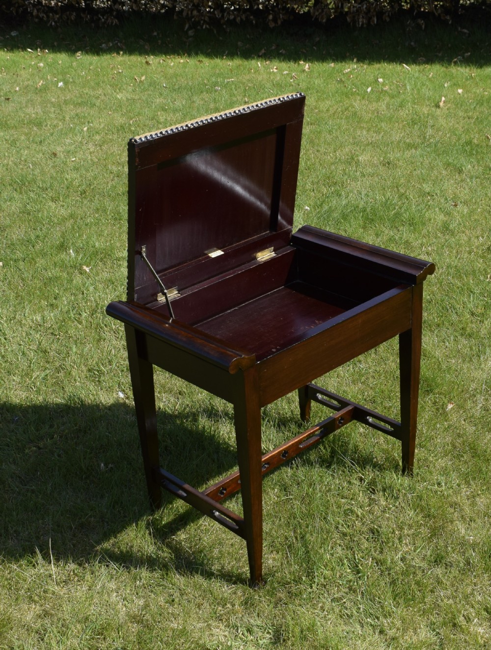 An Arts and Crafts dark wood piano stool featuring a textured tan upholstered seat with brass studding, raised side handles, tapered legs, and a pierced decorative lower stretcher. View IX