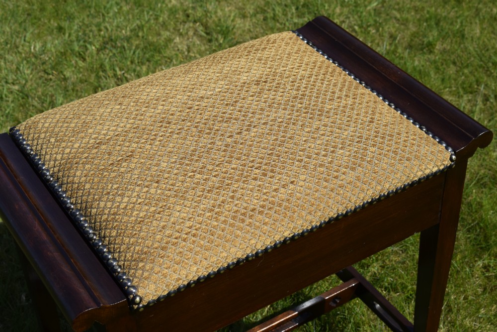 An Arts and Crafts dark wood piano stool featuring a textured tan upholstered seat with brass studding, raised side handles, tapered legs, and a pierced decorative lower stretcher. View XII