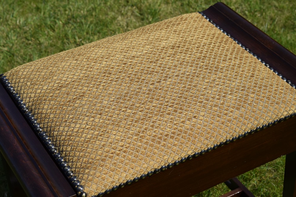 An Arts and Crafts dark wood piano stool featuring a textured tan upholstered seat with brass studding, raised side handles, tapered legs, and a pierced decorative lower stretcher. View XIII