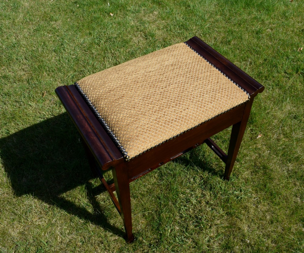 An Arts and Crafts dark wood piano stool featuring a textured tan upholstered seat with brass studding, raised side handles, tapered legs, and a pierced decorative lower stretcher. View XIV