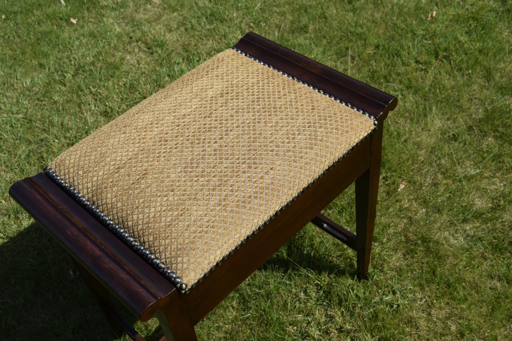 An Arts and Crafts dark wood piano stool featuring a textured tan upholstered seat with brass studding, raised side handles, tapered legs, and a pierced decorative lower stretcher. View XV