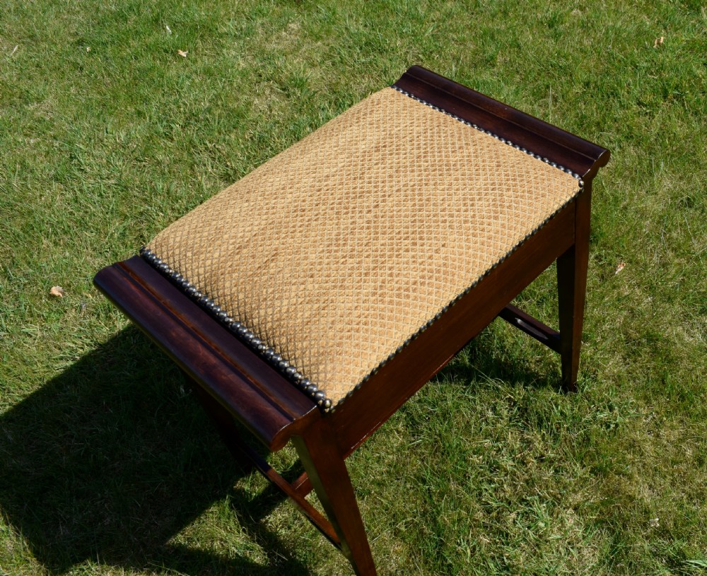 An Arts and Crafts dark wood piano stool featuring a textured tan upholstered seat with brass studding, raised side handles, tapered legs, and a pierced decorative lower stretcher. View XVI