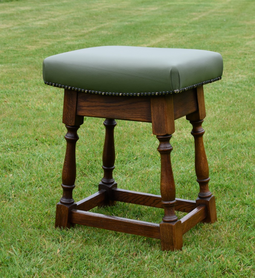 An early twentieth-century oak joint stool featuring an olive green leather upholstered seat secured with brass studs over turned legs. View V