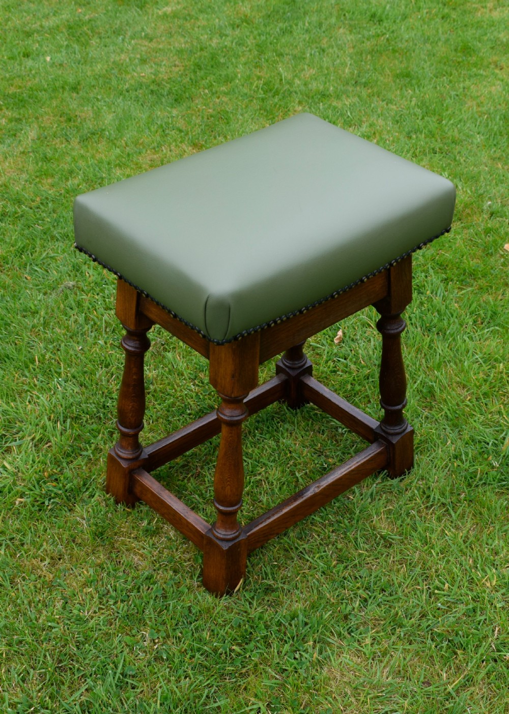 An early twentieth-century oak joint stool featuring an olive green leather upholstered seat secured with brass studs over turned legs. View X