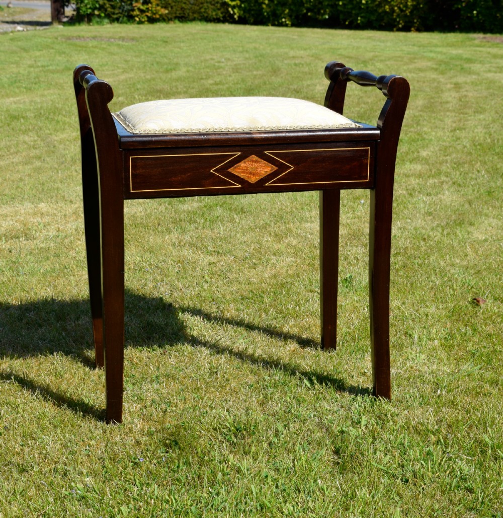 An Edwardian dark mahogany music stool featuring turned side handles, a geometric floral marquetry inlay on the frame, straight tapered legs, and a cream upholstered padded seat.