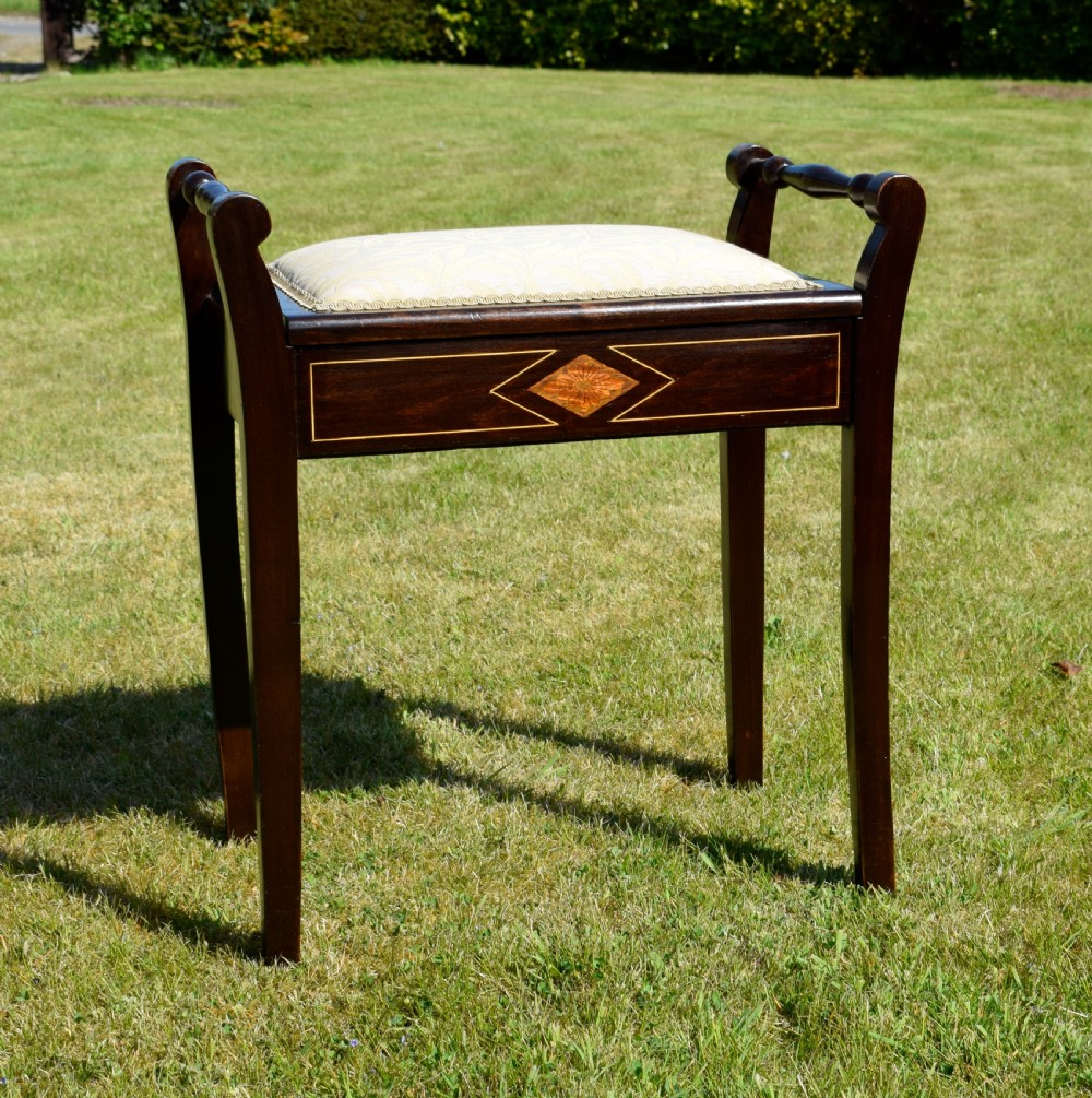 An Edwardian dark mahogany music stool featuring turned side handles, a geometric floral marquetry inlay on the frame, straight tapered legs, and a cream upholstered padded seat. View II