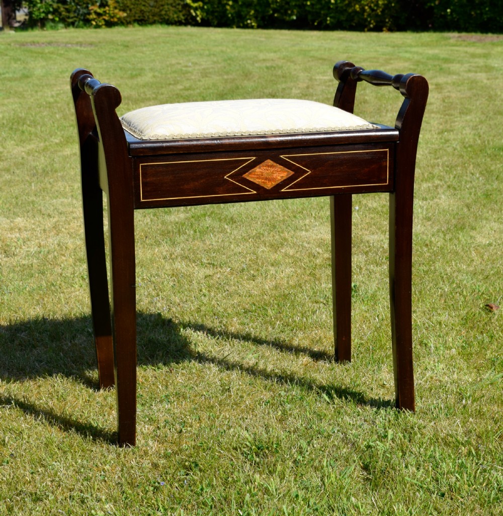 An Edwardian dark mahogany music stool featuring turned side handles, a geometric floral marquetry inlay on the frame, straight tapered legs, and a cream upholstered padded seat. View III