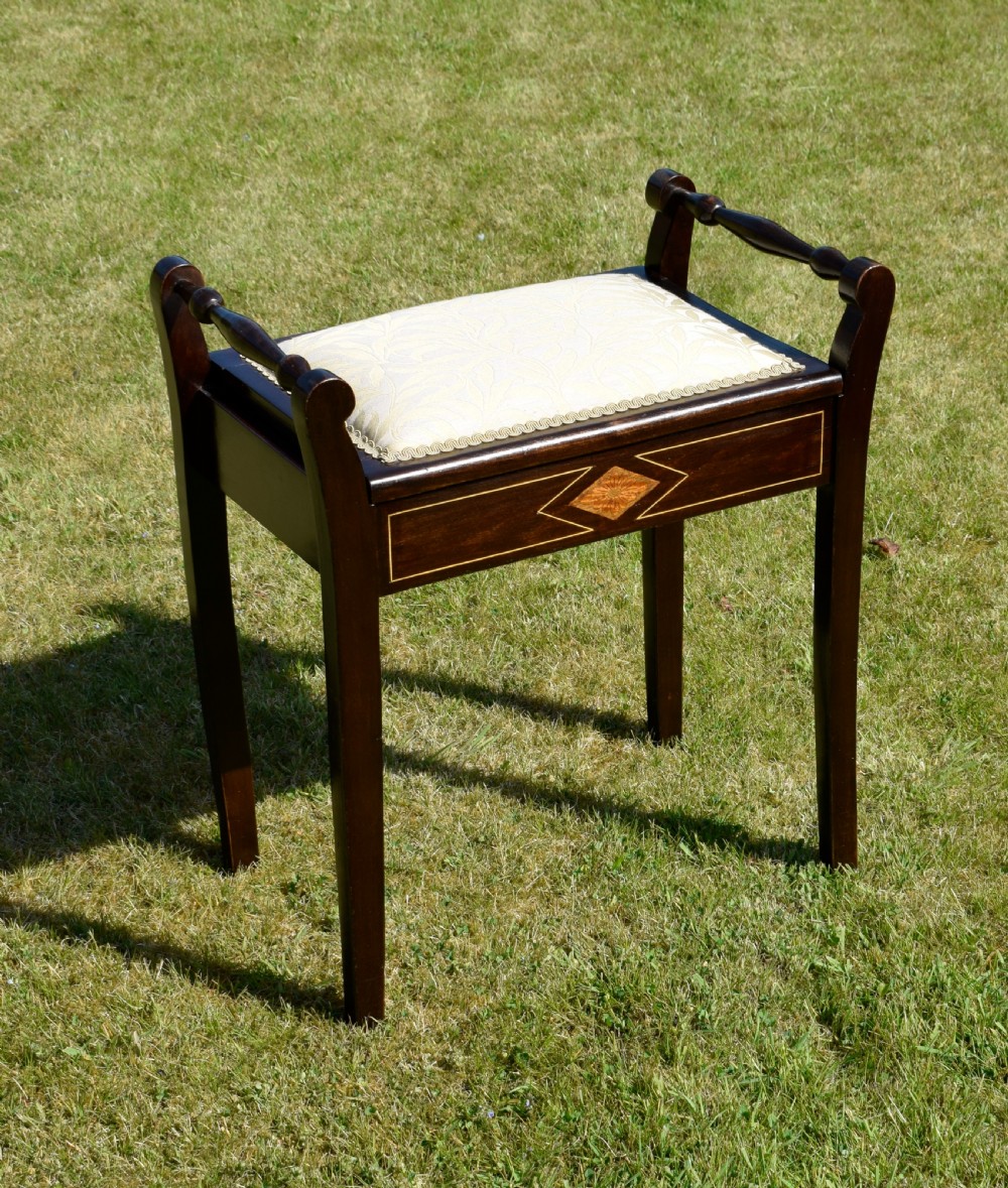 An Edwardian dark mahogany music stool featuring turned side handles, a geometric floral marquetry inlay on the frame, straight tapered legs, and a cream upholstered padded seat. View V