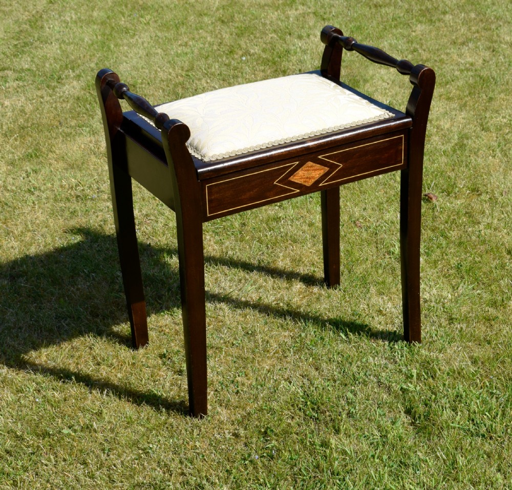An Edwardian dark mahogany music stool featuring turned side handles, a geometric floral marquetry inlay on the frame, straight tapered legs, and a cream upholstered padded seat. View VI