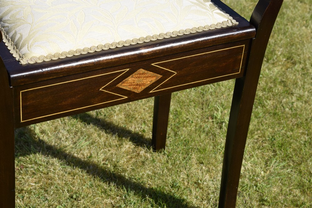 An Edwardian dark mahogany music stool featuring turned side handles, a geometric floral marquetry inlay on the frame, straight tapered legs, and a cream upholstered padded seat. View VII