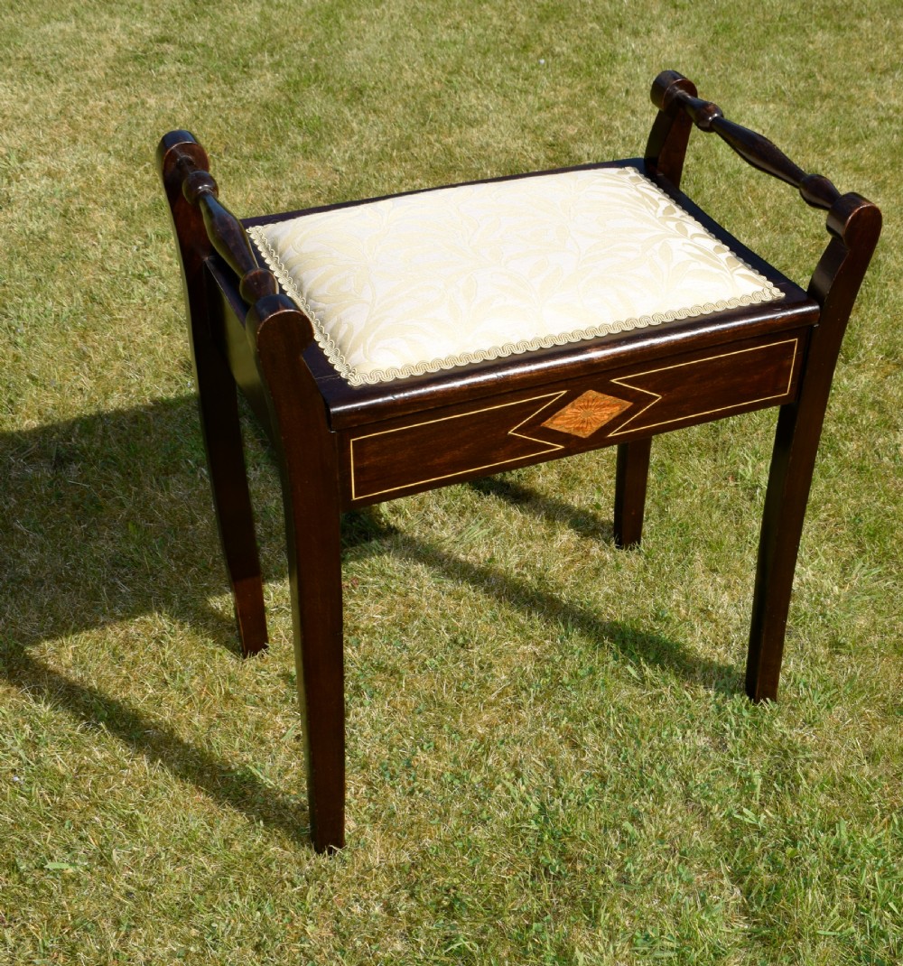 An Edwardian dark mahogany music stool featuring turned side handles, a geometric floral marquetry inlay on the frame, straight tapered legs, and a cream upholstered padded seat. View VIII