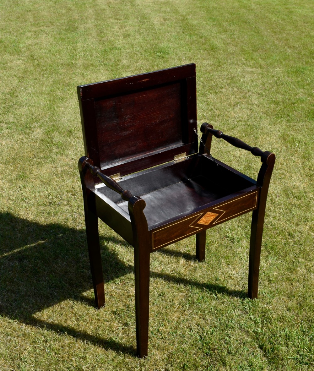 An Edwardian dark mahogany music stool featuring turned side handles, a geometric floral marquetry inlay on the frame, straight tapered legs, and a cream upholstered padded seat. View IX