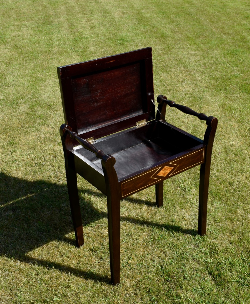 An Edwardian dark mahogany music stool featuring turned side handles, a geometric floral marquetry inlay on the frame, straight tapered legs, and a cream upholstered padded seat. View X