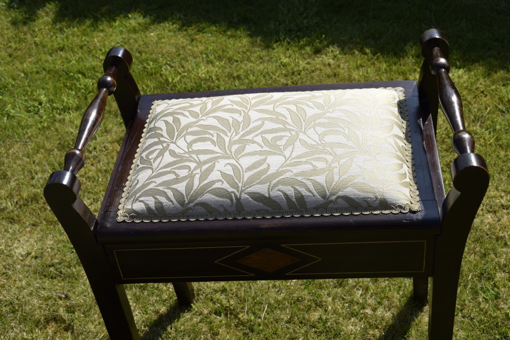 An Edwardian dark mahogany music stool featuring turned side handles, a geometric floral marquetry inlay on the frame, straight tapered legs, and a cream upholstered padded seat. View XV
