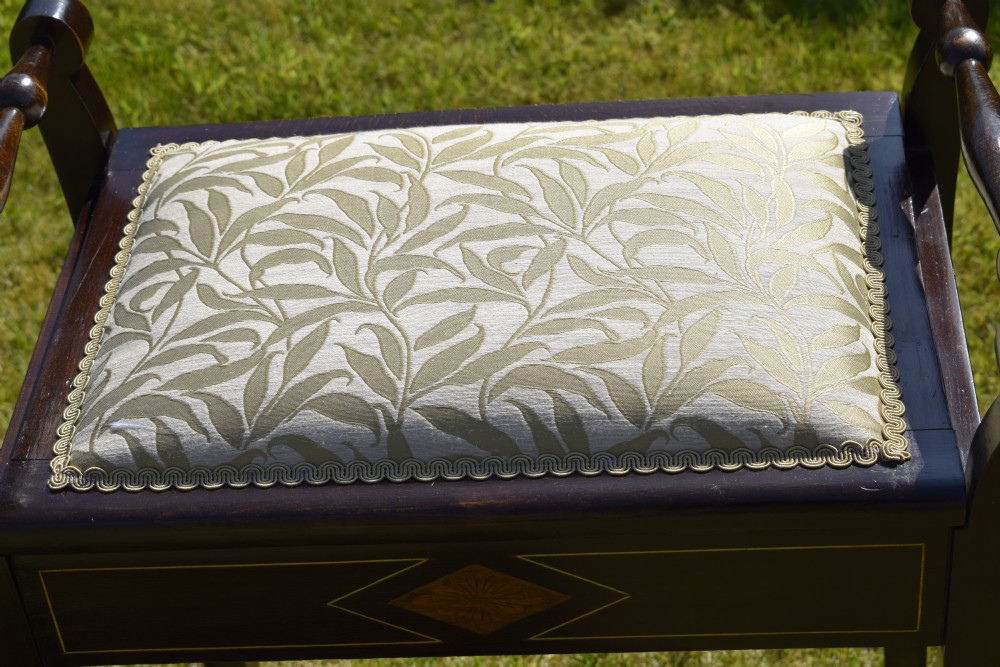 An Edwardian dark mahogany music stool featuring turned side handles, a geometric floral marquetry inlay on the frame, straight tapered legs, and a cream upholstered padded seat. View XVI