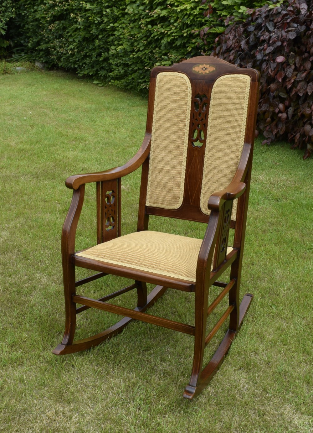 An Edwardian mahogany rocking chair featuring a crest rail with central marquetry inlay, double upholstered back panels, pierced fretwork splats, and light beige textured upholstery.