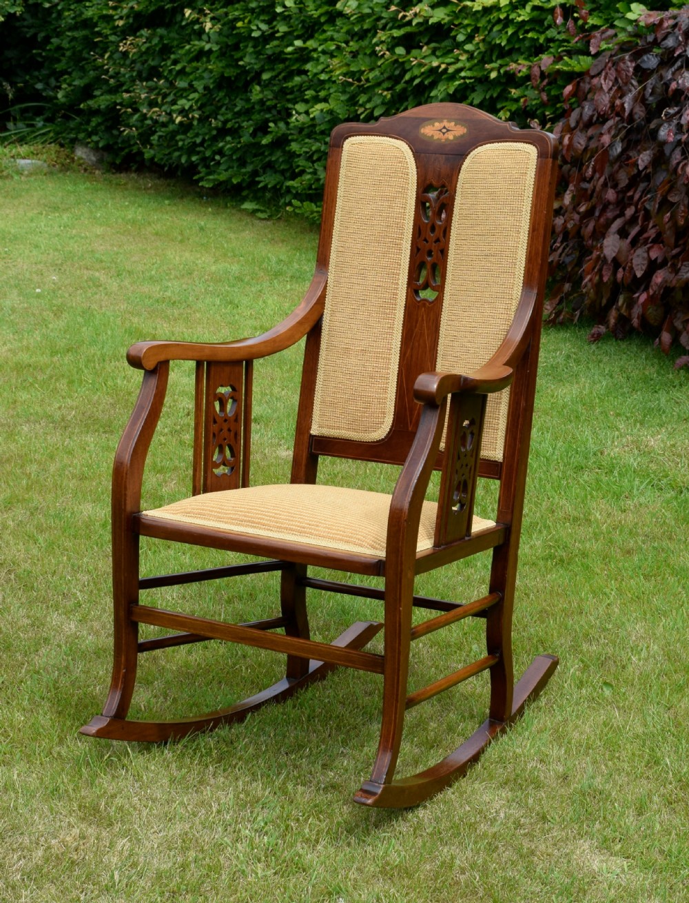 An Edwardian mahogany rocking chair featuring a crest rail with central marquetry inlay, double upholstered back panels, pierced fretwork splats, and light beige textured upholstery. View II