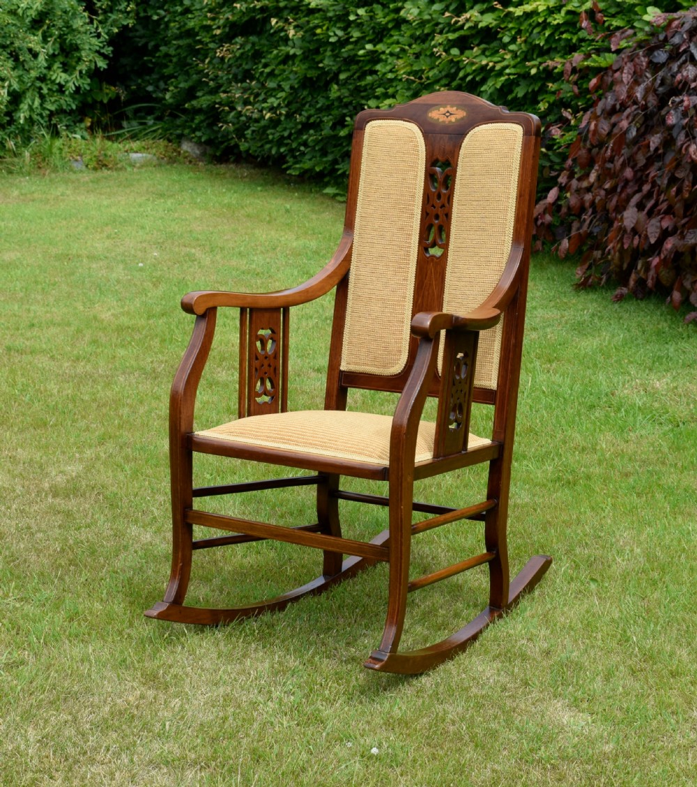 An Edwardian mahogany rocking chair featuring a crest rail with central marquetry inlay, double upholstered back panels, pierced fretwork splats, and light beige textured upholstery. View III