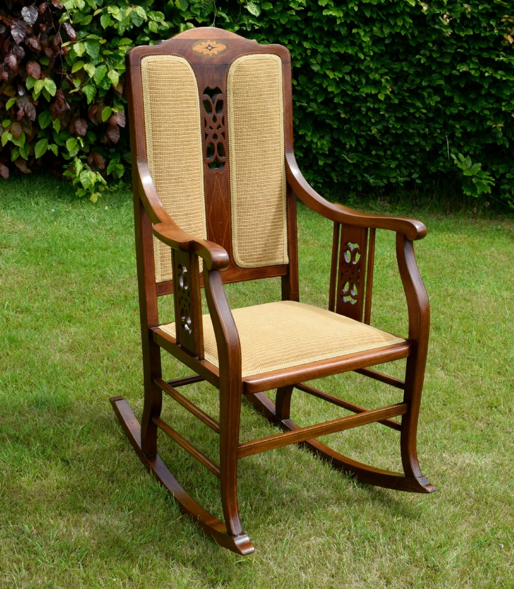 An Edwardian mahogany rocking chair featuring a crest rail with central marquetry inlay, double upholstered back panels, pierced fretwork splats, and light beige textured upholstery. View V