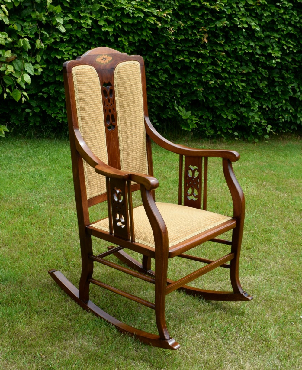 An Edwardian mahogany rocking chair featuring a crest rail with central marquetry inlay, double upholstered back panels, pierced fretwork splats, and light beige textured upholstery. View VI