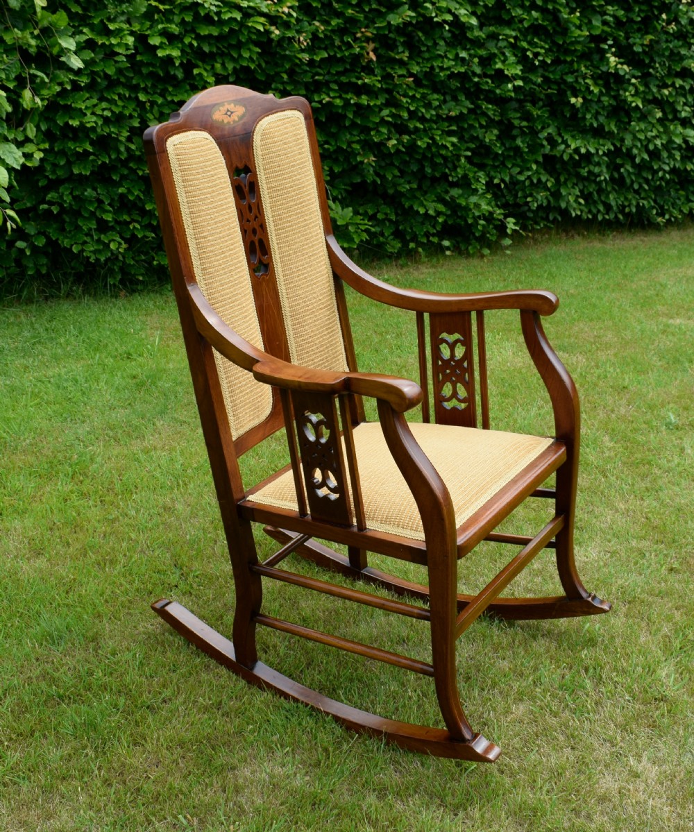 An Edwardian mahogany rocking chair featuring a crest rail with central marquetry inlay, double upholstered back panels, pierced fretwork splats, and light beige textured upholstery. View IX