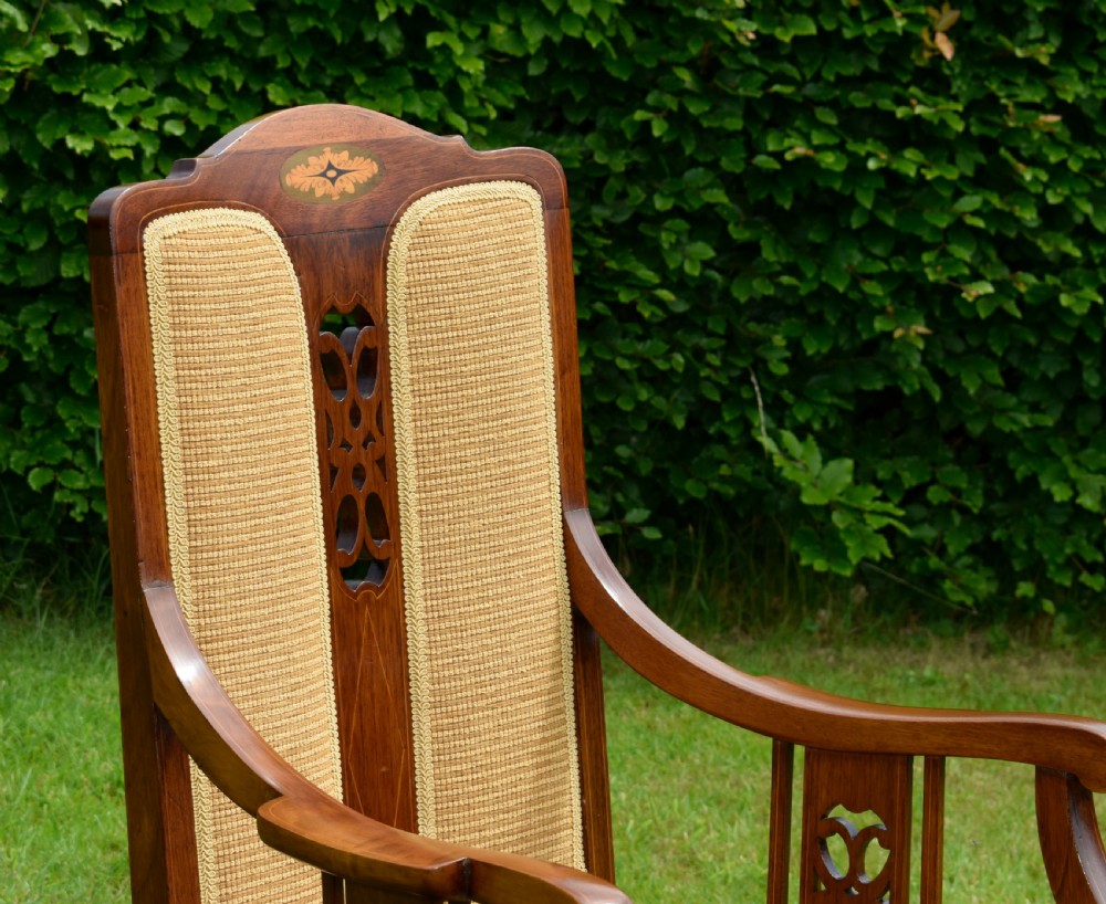 An Edwardian mahogany rocking chair featuring a crest rail with central marquetry inlay, double upholstered back panels, pierced fretwork splats, and light beige textured upholstery. View X
