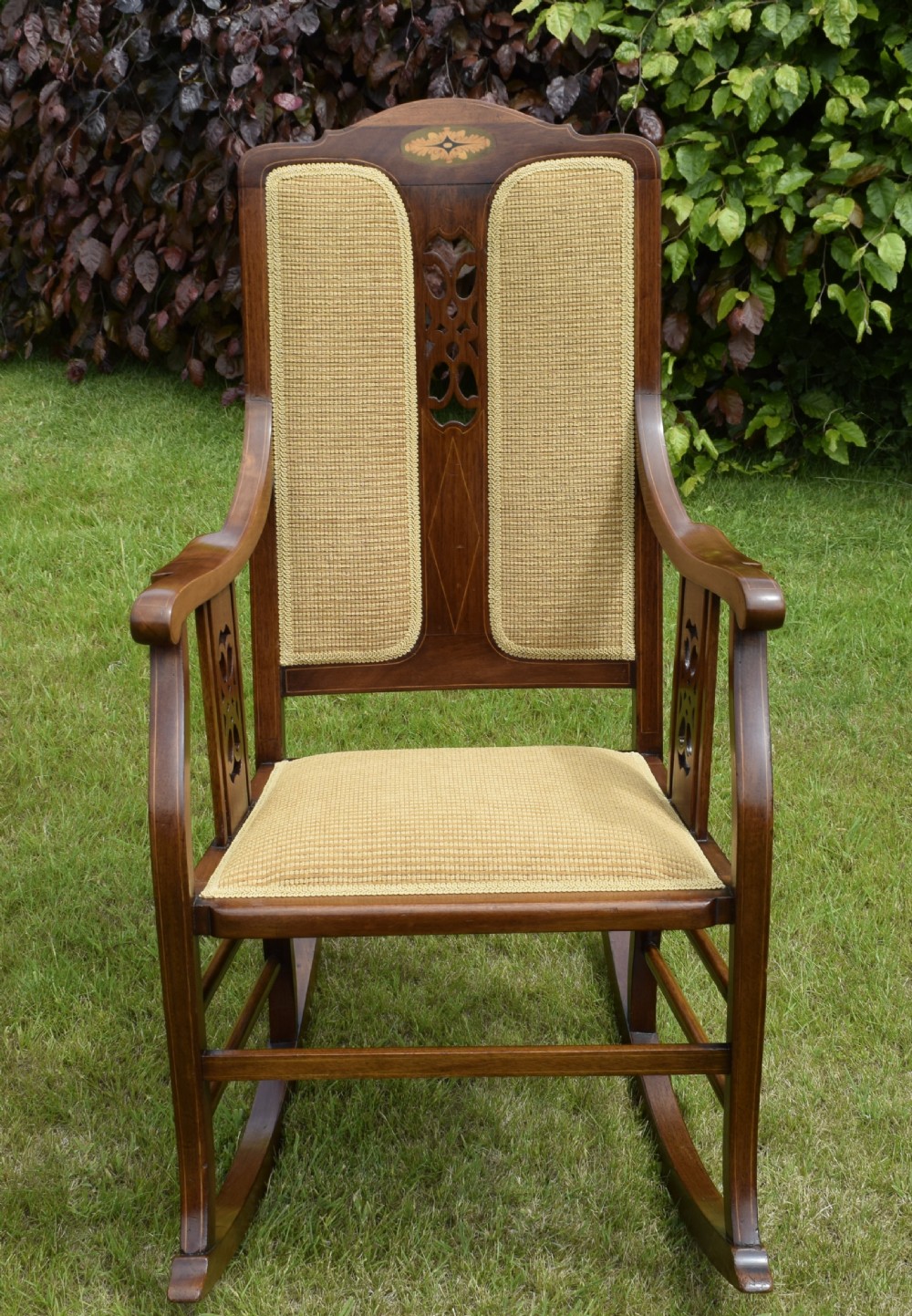 An Edwardian mahogany rocking chair featuring a crest rail with central marquetry inlay, double upholstered back panels, pierced fretwork splats, and light beige textured upholstery. View XI