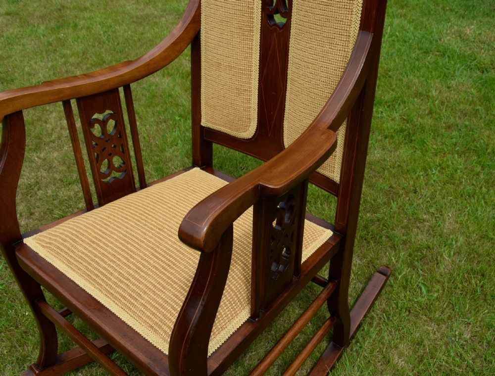 An Edwardian mahogany rocking chair featuring a crest rail with central marquetry inlay, double upholstered back panels, pierced fretwork splats, and light beige textured upholstery. View XIV