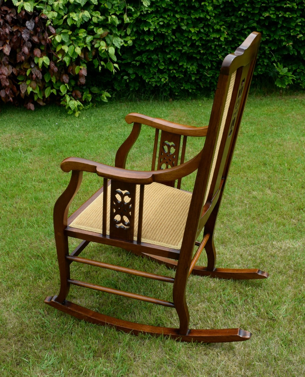 An Edwardian mahogany rocking chair featuring a crest rail with central marquetry inlay, double upholstered back panels, pierced fretwork splats, and light beige textured upholstery. View XV