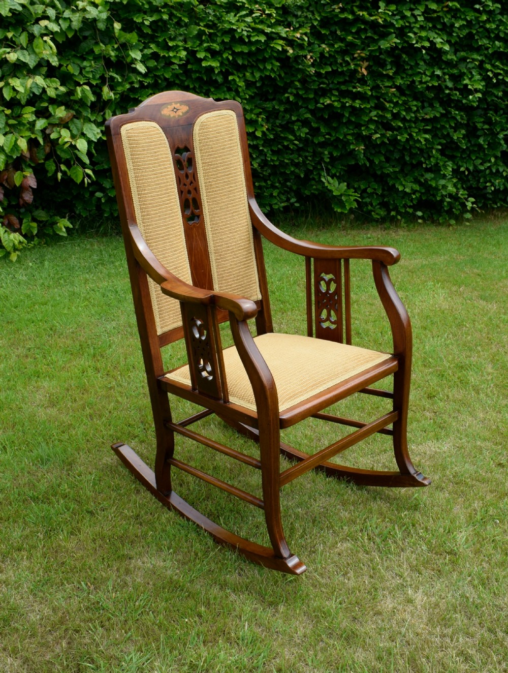 An Edwardian mahogany rocking chair featuring a crest rail with central marquetry inlay, double upholstered back panels, pierced fretwork splats, and light beige textured upholstery. View XVI