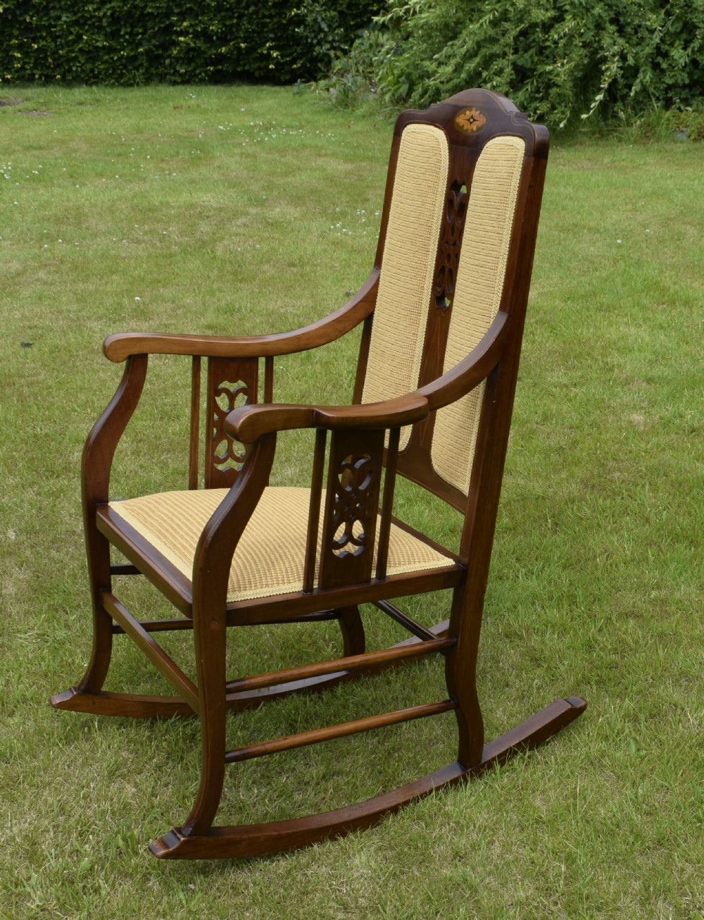 An Edwardian mahogany rocking chair featuring a crest rail with central marquetry inlay, double upholstered back panels, pierced fretwork splats, and light beige textured upholstery. View XVII