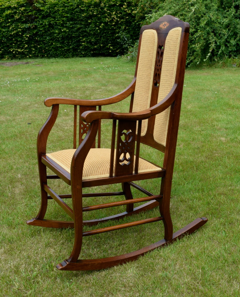 An Edwardian mahogany rocking chair featuring a crest rail with central marquetry inlay, double upholstered back panels, pierced fretwork splats, and light beige textured upholstery. View XVIII