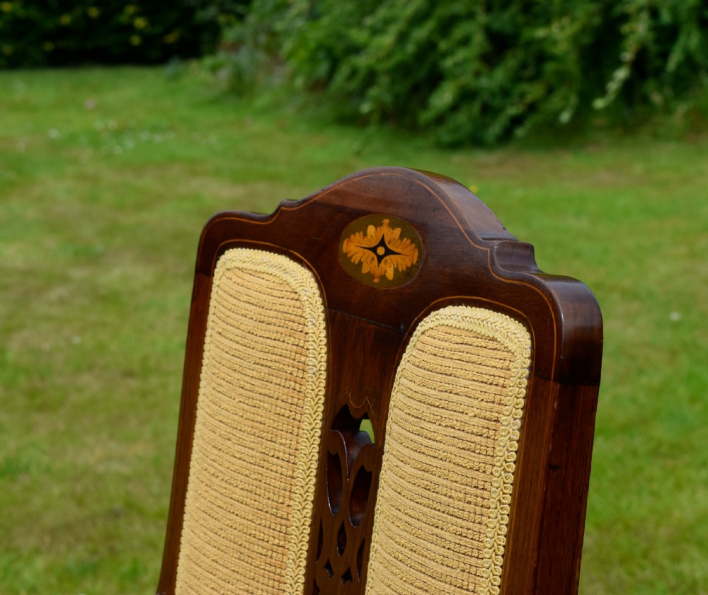 An Edwardian mahogany rocking chair featuring a crest rail with central marquetry inlay, double upholstered back panels, pierced fretwork splats, and light beige textured upholstery. View XIX