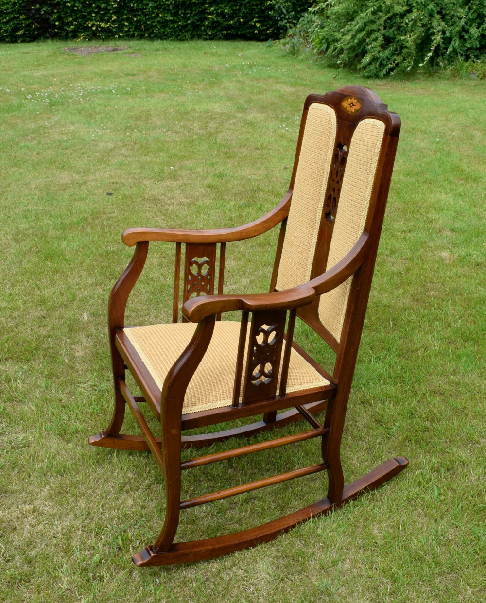 An Edwardian mahogany rocking chair featuring a crest rail with central marquetry inlay, double upholstered back panels, pierced fretwork splats, and light beige textured upholstery. View XXI