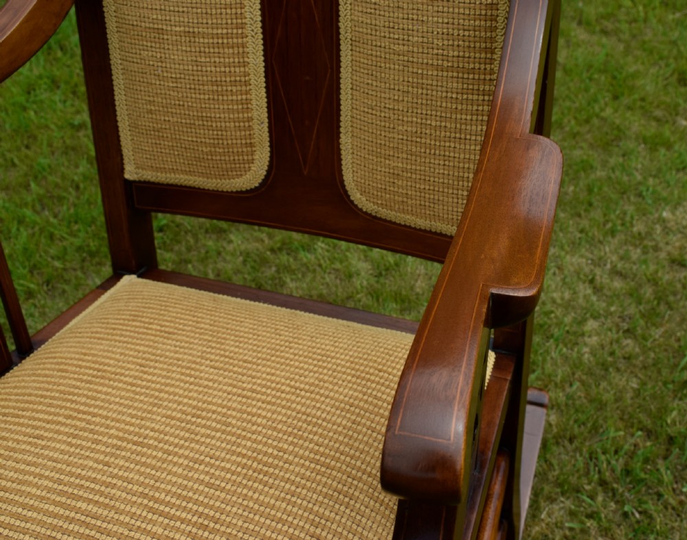 An Edwardian mahogany rocking chair featuring a crest rail with central marquetry inlay, double upholstered back panels, pierced fretwork splats, and light beige textured upholstery. View XXII