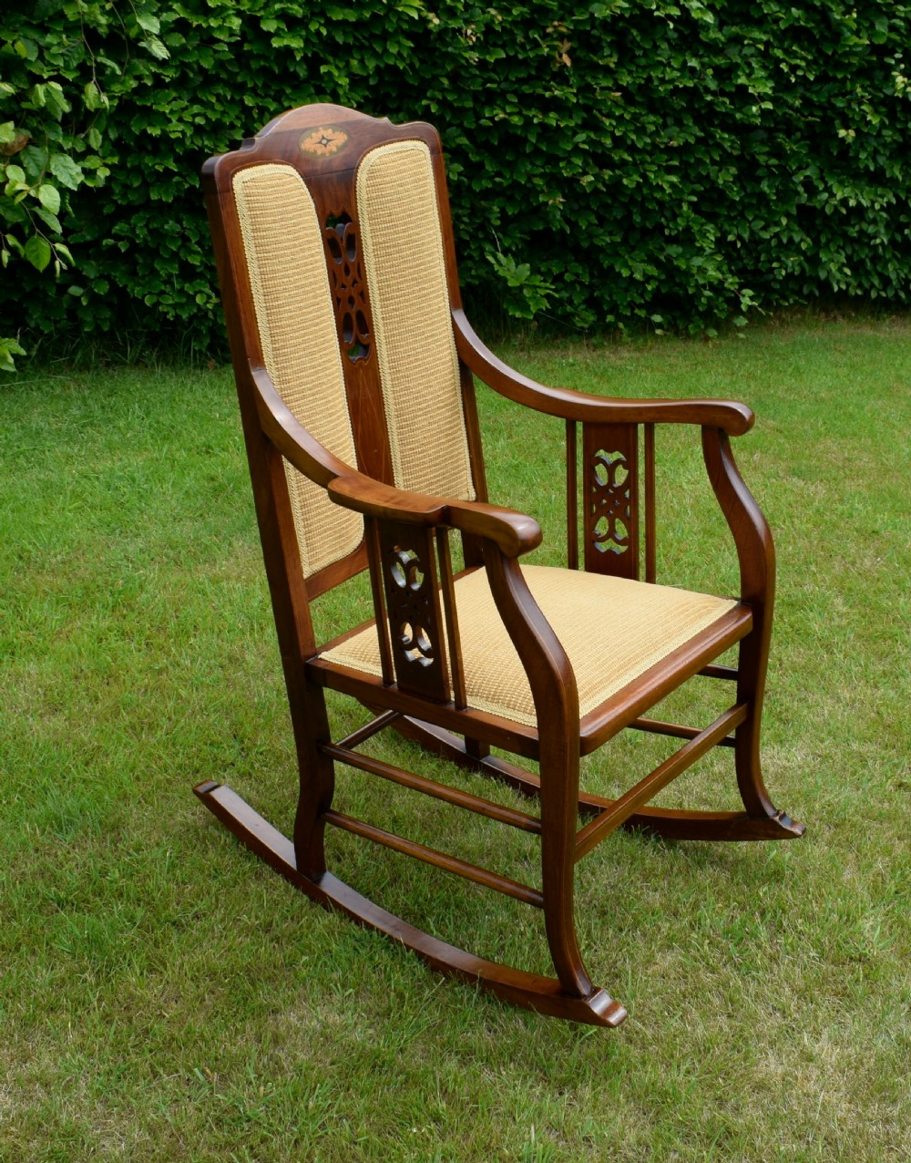 An Edwardian mahogany rocking chair featuring a crest rail with central marquetry inlay, double upholstered back panels, pierced fretwork splats, and light beige textured upholstery. View XXIV